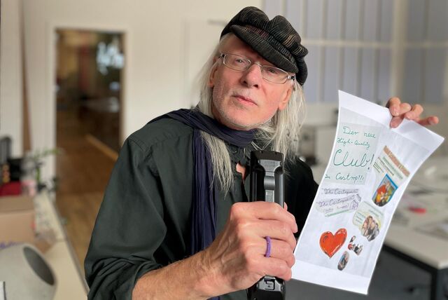 Ein Mann mit Hut namens Rudolf Schiffmann zeigt ein handgeschriebenes Plakat, mit dem er für Konzerte wirbt.