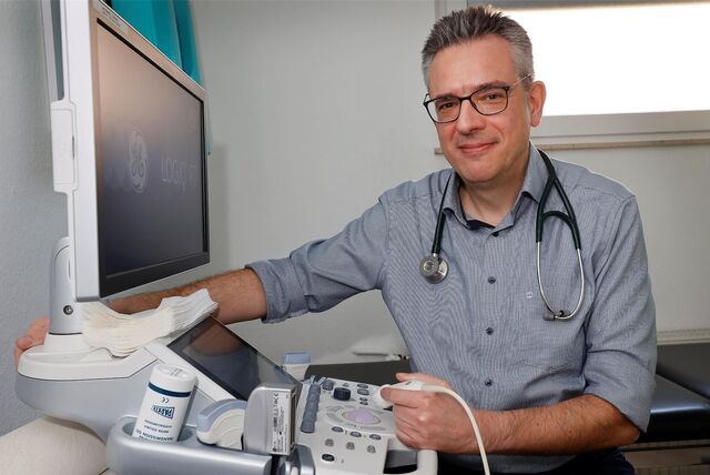 Ein Arzt in weiß-blau-kariertem Hemd sitzt an einem Ultraschallgerät. Er trägt ein Stethoskop um den Hals.