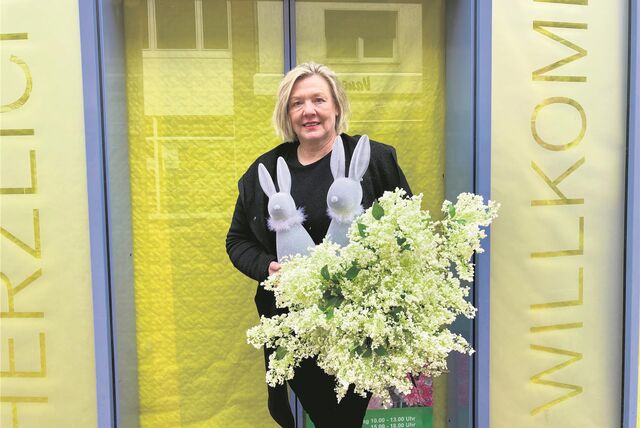 Eine Frau mit zwei Deko-Hasen und Fliederstrauß vor dem zugehängten Deko-Laden.