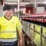 Ein Mitarbeiter von Coca-Cola steht vor einem Fließband mit Coca-Cola-Flaschen.