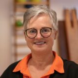 Eine lächelnde Frau mit kurzen, grauen Haaren und schwarzer Brille.