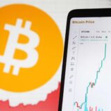 Ein Smartphone, auf dem eine Grafik die Bitcoin-Entwicklung darstellt. Im Hintergrund ist das Bitcoin-Logo zu sehen.
