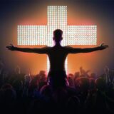 Ein Mann, der Jesus bei der Passion darstellt, steht mit ausgestreckten Armen vor einem hell erleuchteten Kreuz und einer Menschenmenge.