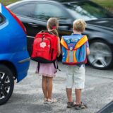Ein Junge und ein Mädchen stehen zwischen parkenden Autos am Straßenrand, während ein Auto vorbeifährt.