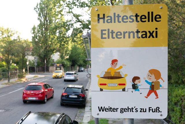 An einer Straße steht ein Schild mit der Aufschrift: „Haltestelle Elterntaxi – weiter geht*s zu Fuß“