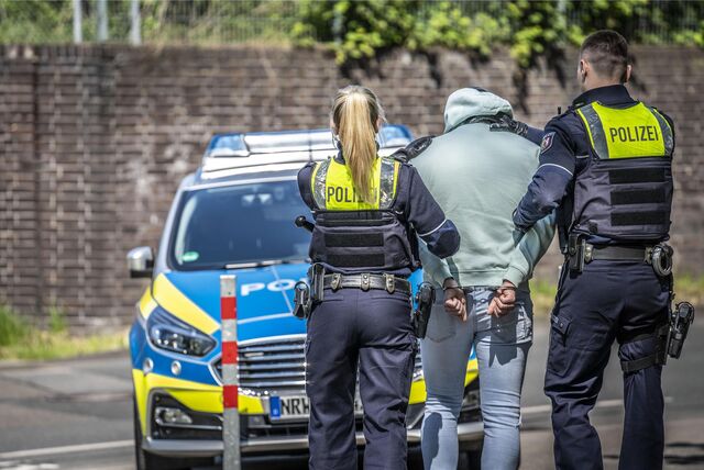 Ein Polizist und eine Polizistin führen einen gefesselten Mann zum Streifenwagen.