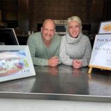 Das Betreiberpaar Marco Pape und Sabrina Macher schauen über die Theke des Drügen Pütt in Haltern, auf der Hinweise auf Matjesbrötchen sowie Kaffee und Kakao stehen.