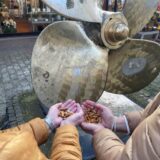 Zwei Personen, deren Gesichter nicht zu erkennen sind, halten in ihren Händen Zigarettenkippen.  Sie stehen vor dem großen Schiffspropeller in der Dattelner Fußgängerzone.