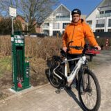 Patrick Berner steht in einem Wohngebiet. Vor sich steht ein weißes Pedelec Fahrrad von KTM. Berner trägt eine orangene Regencke und einen schwarzen Fahrradhelm. Er hält das Fahrrad an Sattel und Lenker. Hinter ihm ist eine Hecke, sowie eine kleine Ansammlung an Büschen. Links neben ihm, steht ein kleiner Service Punkt für Fahrräder und Luftpumpen.