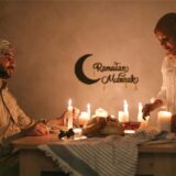 Ein Muslim sitzt an einem mit Kerzen erleuchteten Tisch, während eine Muslima Speisen für das Fastenbrechen (Iftar) während des Ramadan drauf stellt.