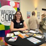Eine Frau steht an einem Stehtisch, hinter ihr hängt ein buntes Werbebanner mit der Aufschrift „Waltrops Dritter Ort“. Im Hintergrund sind Besucher der Freizeitmesse zu erkennen.