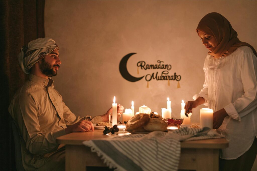 Ein Muslim sitzt an einem mit Kerzen erleuchteten Tisch, während eine Muslima Speisen für das Fastenbrechen (Iftar) während des Ramadan drauf stellt.