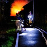 Zwei Feuerwehrmänner auf einer Straße. Im Hintergrund ist Feuerschein am Himmel zu sehen.