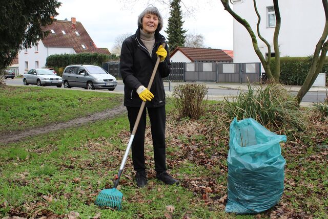 Eine Frau steht mit einer Harke auf einer Rasenfläche, neben ihr ein blauer Müllsack.
