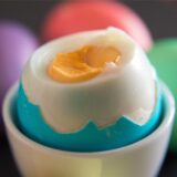 Ein gekochtes und aufgeschlagenes blaues Osterei ist in einem Eierbecher zu sehen. Im Hintergrund liegen drei weitere bunte Ostereier.