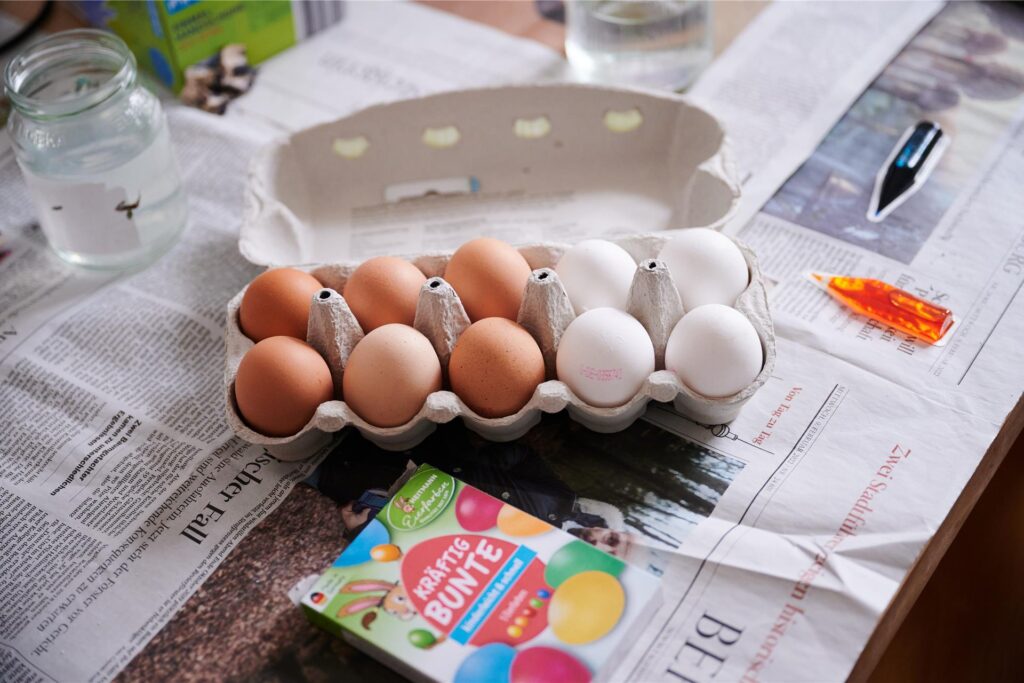 Ein Eierkarton mit sechs braunen und vier weißen Eiern steht auf einem Tisch, der mit Zeitungspaper ausgelegt ist. Daneben liegt ein Paket mit Farben für Ostereier. Ein Eierkarton mit sechs braunen und vier weißen Eiern steht auf einem Tisch, der mit Zeitungspaper ausgelegt ist. Daneben liegt ein Paket mit Farben für Ostereier.