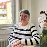 Monika Verhoven sitzt an einem Tisch, neben ihr stehen ein paar Kunstblumen in einer grünen Vase.