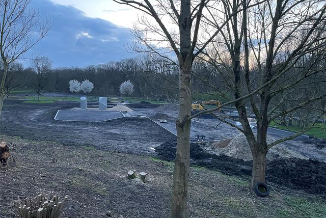 Hinter dem Hallenbad läuft der zweite Bauabschnitt zum Spiel-, Sport- und Bewegungspark. Die Boulderfelsen stehen schon. Vor der Baustelle sind Bäume zu sehen. Hinten blühen drei weitere Bäume.