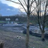 Hinter dem Hallenbad läuft der zweite Bauabschnitt zum Spiel-, Sport- und Bewegungspark. Die Boulderfelsen stehen schon. Vor der Baustelle sind Bäume zu sehen. Hinten blühen drei weitere Bäume.