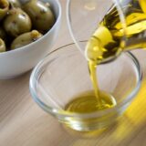 Olivenöl wird in eine Glasschale gefüllt, während im Hintergrund eine Schale mit grünen Oliven steht.
