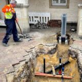 Ein Gelsenwasser-Mitarbeiter legt mit einer Schippe die aufgenommenen Pflastersteine zusammen. Daneben ist der Brunnen zu sehen.