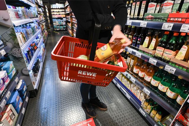 Eine Frau steht bei Rewe vor einem Lebensmittelregal und legt eine Flasche Speiseöl in ihren Einkaufskorb.