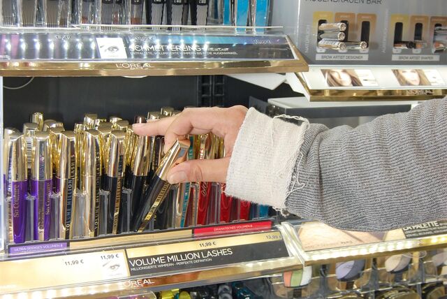 Auf dem Symbolfoto zum Thema Ladendiebstahl ist ein Frauenarm, bedeckt mit einem grau-beigen Ärmel, zu sehen sowie die Hand einer Frau. Diese greift nach einer Mascara in einem Regal voller Kosmetik.