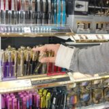 Auf dem Symbolfoto zum Thema Ladendiebstahl ist ein Frauenarm, bedeckt mit einem grau-beigen Ärmel, zu sehen sowie die Hand einer Frau. Diese greift nach einer Mascara in einem Regal voller Kosmetik.