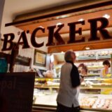 „Bäckerei“ ist am Schaufenster einer Bäckerei zu lesen, in der ein Kunde von zwei Mitarbeitern bedient wird.