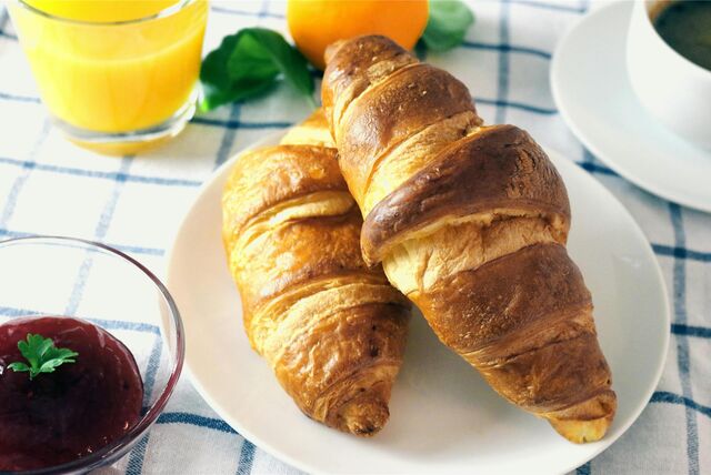 Ein Teller mit zwei Croissants steht zusammen mit einem Glas Orangensaft, einer Tasse Kaffee und einer Schale Marmelade auf einem Frühstückstisch.