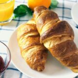 Ein Teller mit zwei Croissants steht zusammen mit einem Glas Orangensaft, einer Tasse Kaffee und einer Schale Marmelade auf einem Frühstückstisch.