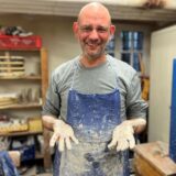 Dirk Wenke trägt eine blaue Schürze und hat Ton an den Händen. Er lächelt in die Kamera.