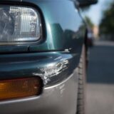 An der Vorderschürze zerstörtes Auto, das parkt, daneben Blick auf eine lange Straße.