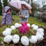 Zwei Mädchen stehen in einem Garten, eine hält einen Regenschirm und ein Osternest in den Händen, im Vordergrund sieht man einen rosafarbenen Deko-Hasen und weiße Eier.