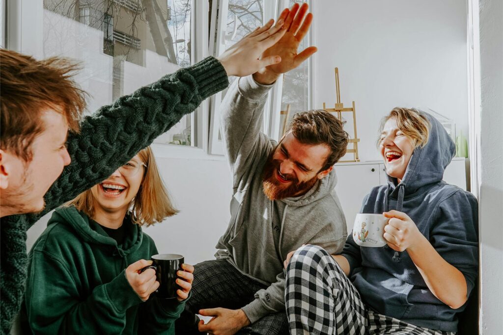 Vier Personen sitzen zusammen, trinken Kaffee, lachen und zwei von ihnen geben sich ein High five. Vier Personen sitzen zusammen, trinken Kaffee, lachen und zwei von ihnen geben sich ein High five.