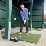 Ein Mann steht in einem halbseitig geöffnete Holz-Haus auf einem grünen Teppich-Quadrat, auf dem ein Golfball liegt. Er hat einen Schläger in der Hand, um gleich den Abschlag zu üben.