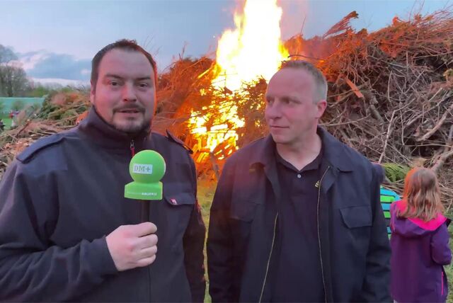 Zwei Männer stehen mit einem Mikrofon vor einem brennenden Osterfeuer.