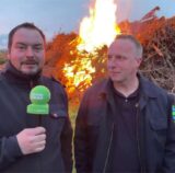Zwei Männer stehen mit einem Mikrofon vor einem brennenden Osterfeuer.