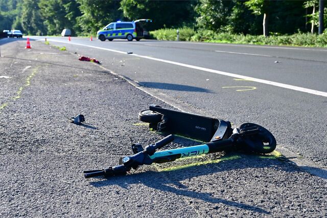 Ein schwer beschädigter E-Scooter liegt auf einer Straße, im Hintergrund ein Polizei-Fahrzeug.