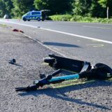 Ein schwer beschädigter E-Scooter liegt auf einer Straße, im Hintergrund ein Polizei-Fahrzeug.