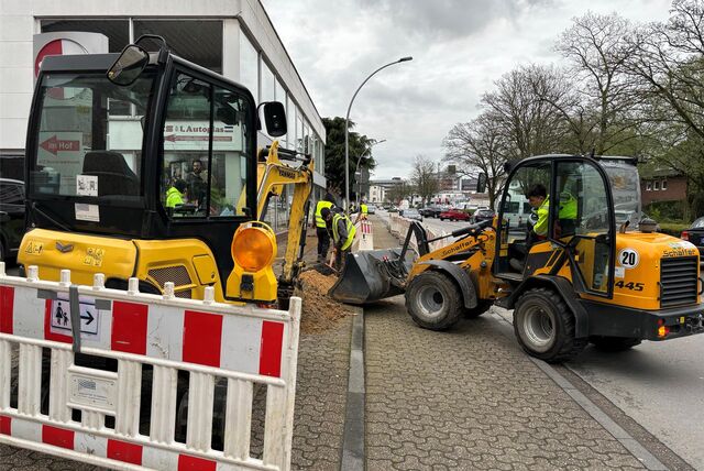Ansicht einer Baustelle auf einem Gehweg. Links im Vordergrund ist eine Baustellenbake zu erkennen. Rechts im Bild steht ein Bagger.