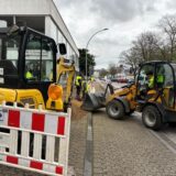 Ansicht einer Baustelle auf einem Gehweg. Links im Vordergrund ist eine Baustellenbake zu erkennen. Rechts im Bild steht ein Bagger.