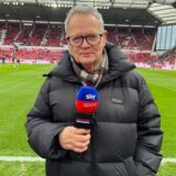 Ein Sportreporter steht im Stadion und hält ein Mikro in der Hand.