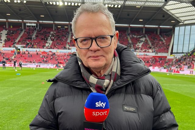 Ein Sportreporter steht im Stadion und hält ein Mikro in der Hand.