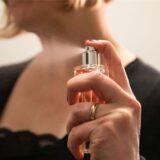 Das Symbolfoto zeigt eine weibliche Hand mit einem Parfum und einer Frau im Hintergrund.