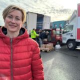 Eine Frau steht vor einem Lkw, der mit Paletten voller Lebensmittel beladen wird. Im Hintergrund sind Männer zu erkennen, die Paletten in ein Fahrzeug laden.