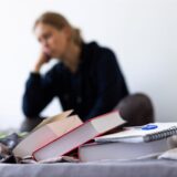 Eine Studentin sitzt auf dem Bett in ihrem WG-Zimmer, vor ihr liegen Lehrbücher und ein Notizbuch.