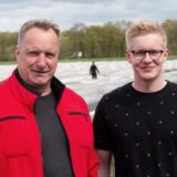 Zwei Männer stehen vor einem Spargelfeld, im Hintergrund arbeiten Erntehelfer.