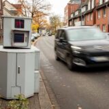 Ein hellgrauer Panzerblitzer der Stadt Herten steht am Fahrbahnrand der Kurt-Schumacher-Straße. Neben ihm fährt gerade ein schwarzer Citroen-Kombi vorbei.