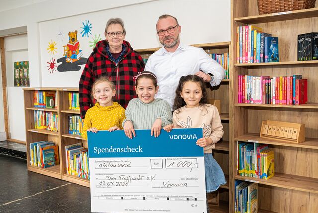 Fünf Personen stehen vor einem Bücherregal und halten einen großen Spendenscheck in der Hand. Sie haben eine Bücherei neu eröffnet.